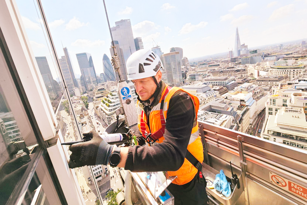 Construction worker in safety gear caulking a high-rise window with London cityscape in the background.