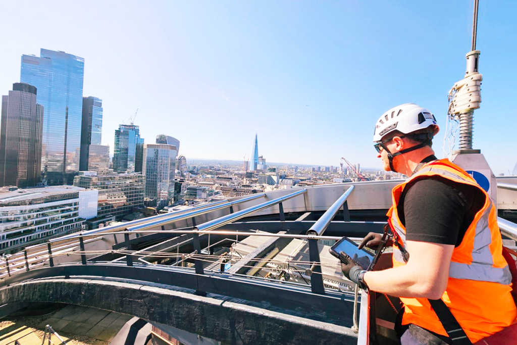Construction worker in orange vest and helmet checks tablet high above London skyline.