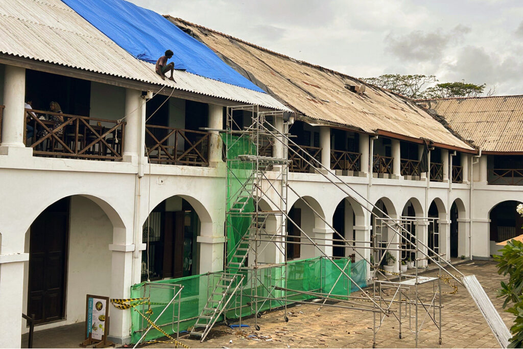 Dilapidated building with blue tarp on roof, scaffolding, and people on terrace; cloudy sky overhead.