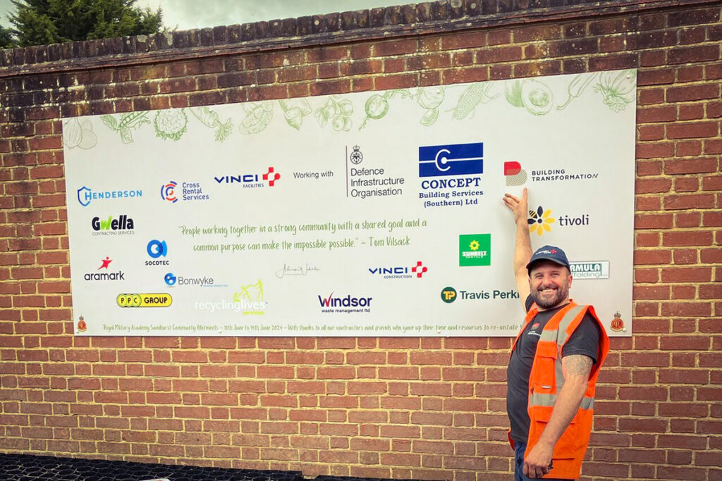 Smiling worker in orange vest points at a sponsor board on brick wall, featuring the Building transformation logo.