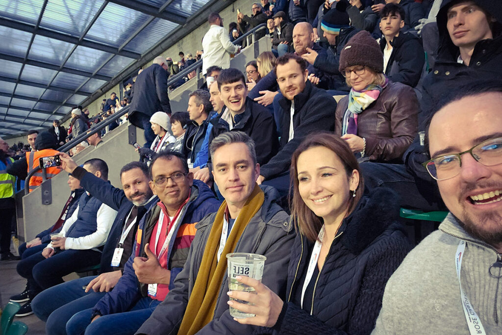 Cheerful group of Building Transformation staff in stadium seats with a crowded background.