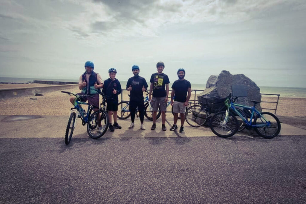 Five cyclists in helmets giving thumbs up by the seaside on a cloudy day, bikes resting beside a large rock.