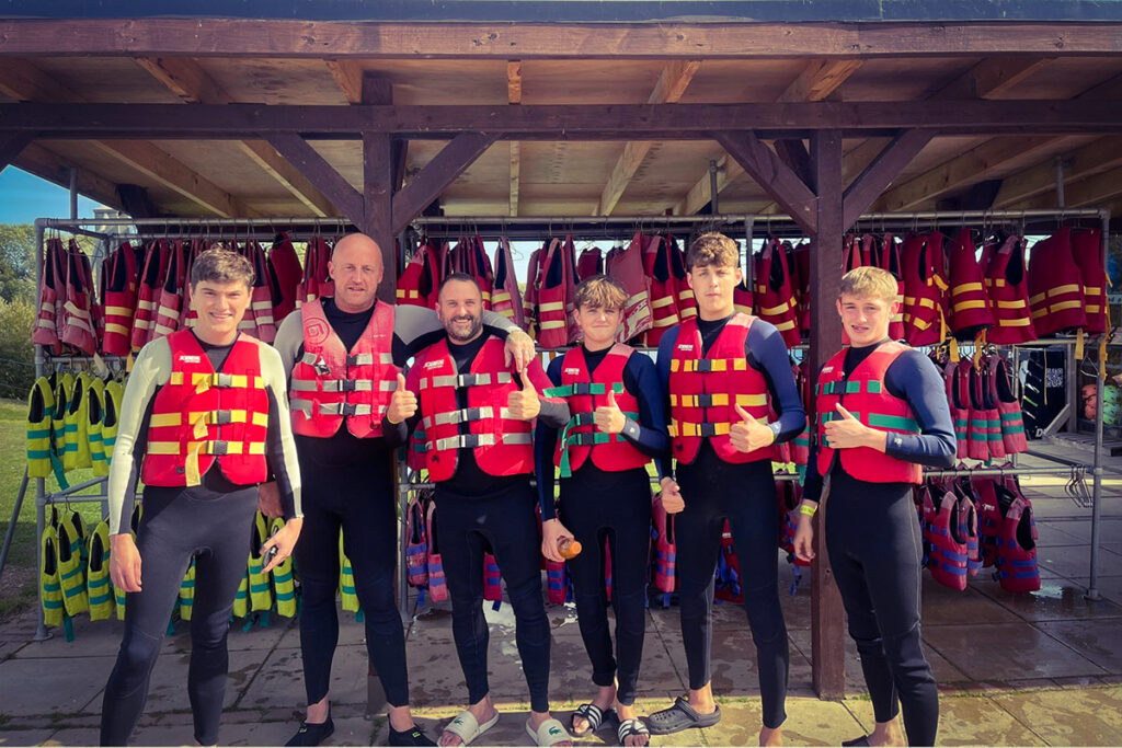 Group of six people in wetsuits and red life vests, giving thumbs up under outdoor shelter.