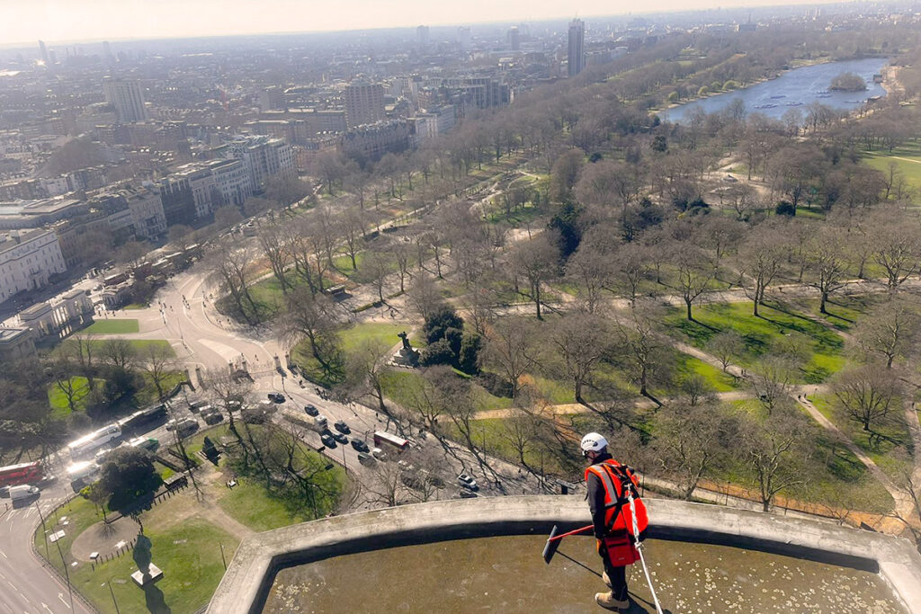 Worker in high-visibility gear conduct electronic leak detection on a rooftop overlooking expansive London park and cityscape.