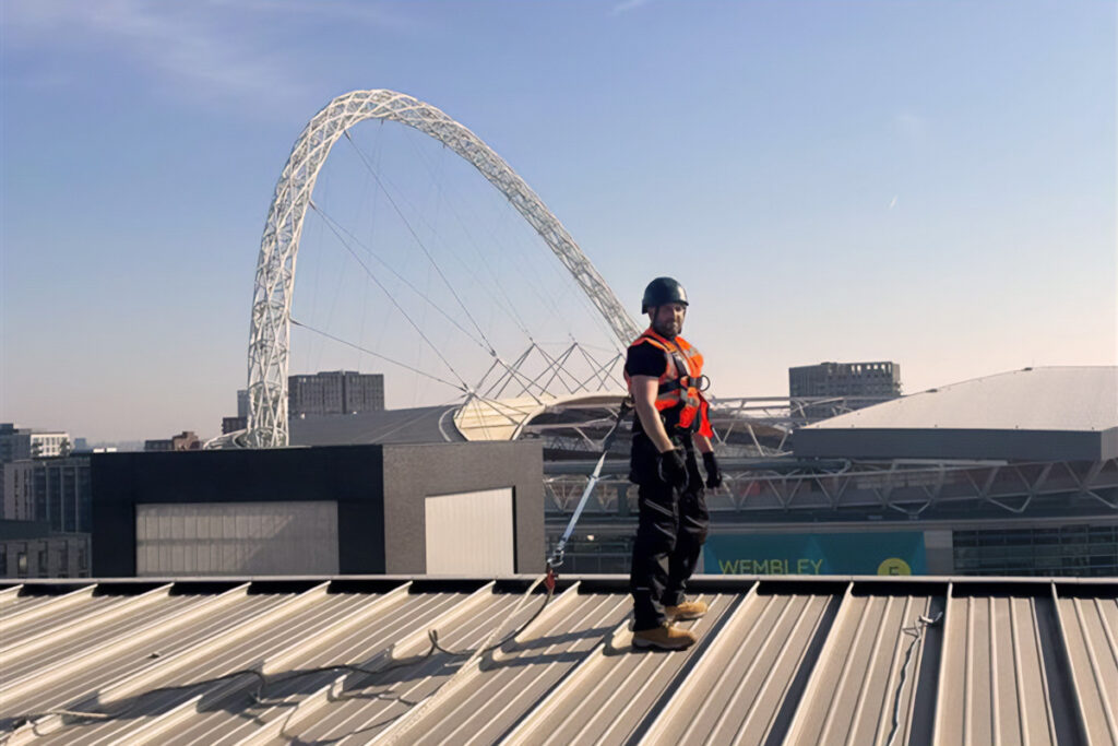 Construction worker with safety gear stands on a building roof near Wembley Stadium arch in London.