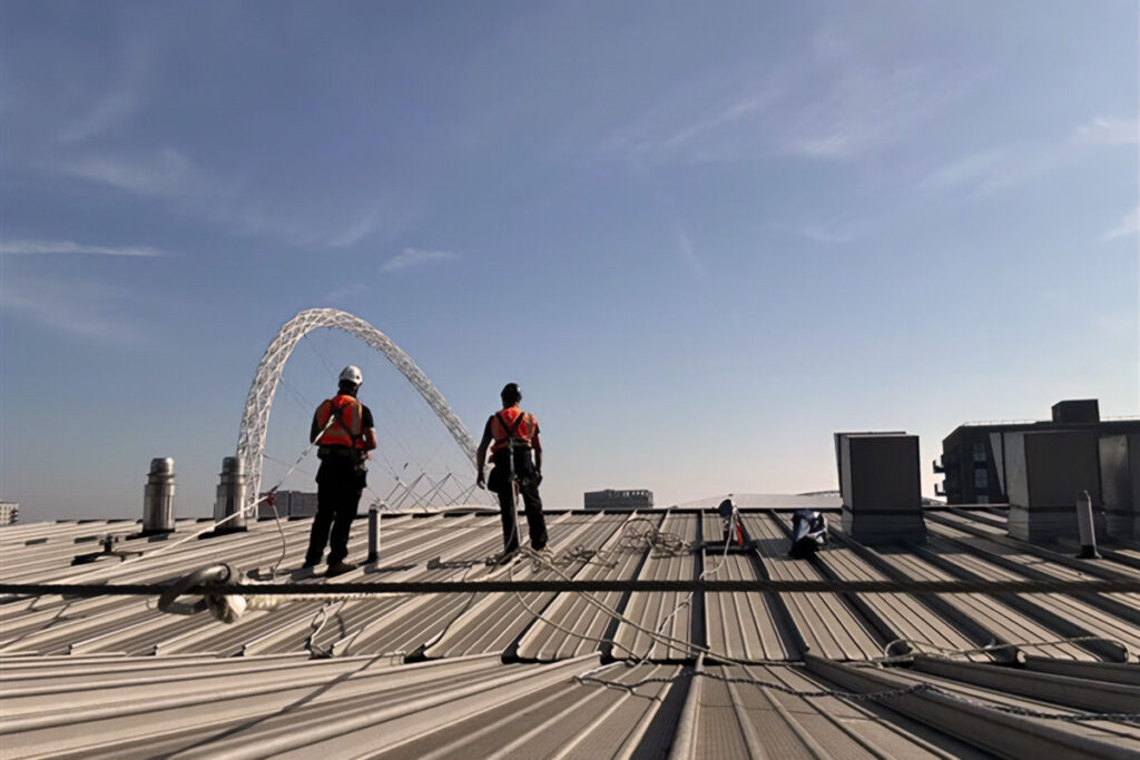 Construction workers with safety gear inspect a rooftop near Wembley Stadium arch under clear skies.