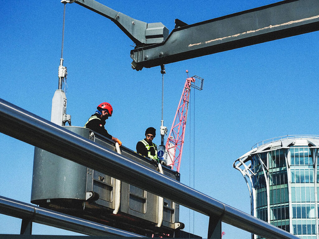 Construction workers in a crane basket with vibrant blue sky, red crane, and modern glass building.