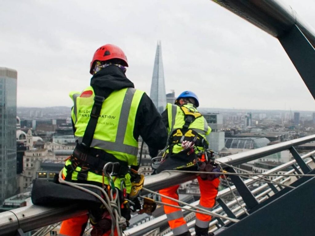 Construction workers in safety gear atop a skyscraper in London, with the Shard visible in the background.