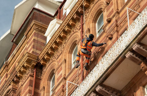 Worker in high-vis gear abseiling down a historic brick building, touching ornate white ironwork.