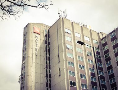Tall ibis hotel building with a beige facade, urban backdrop, and cloudy sky, framed by tree branches.