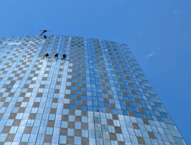 Window cleaners suspended on ropes against a reflective skyscraper facade under a clear blue sky.