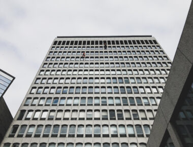 Worker suspended cleaning large office building windows against overcast sky.