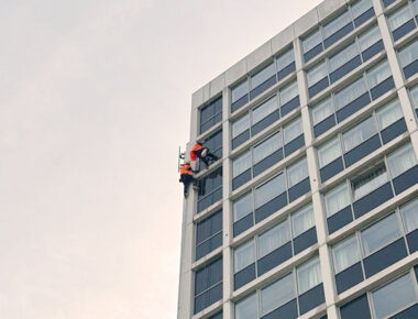 Window cleaner in red uniform scales a tall, modern building using a harness, against a grey sky.