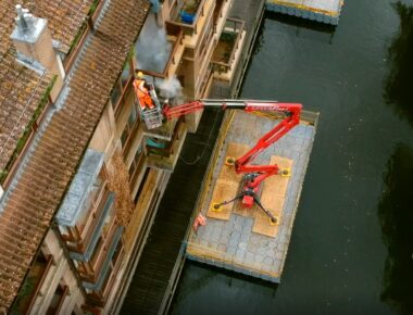 Worker in an orange suit operates a red Spiderlift platform over water near a building, performing maintenance.