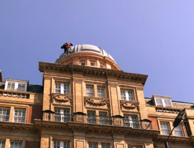 Worker in harness atop ornate brick building with domed roof under clear blue sky.