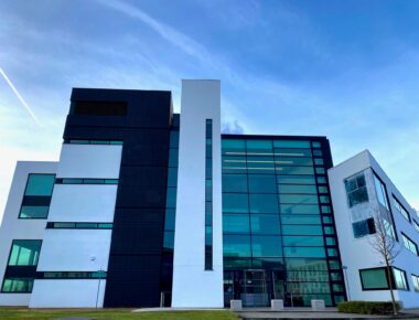 Modern glass office building with angular architecture, set against a vibrant blue sky with a streaking contrail.